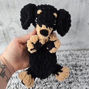 May include: A crocheted plush toy dachshund with black and tan fur. The toy is sitting on a fluffy white rug.