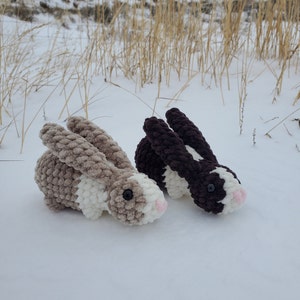 May include: Two crocheted bunny rabbits, one brown and white and one gray and white, are lying on a snowy surface.