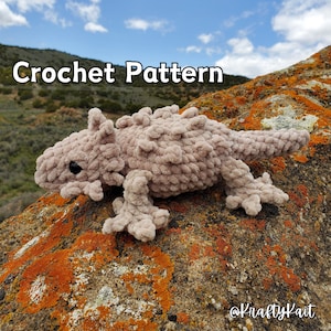 May include: Crochet pattern for a beige crocheted lizard with a spiky back, sitting on a rock with orange lichen. The pattern is available on Etsy, @KraftyKait.