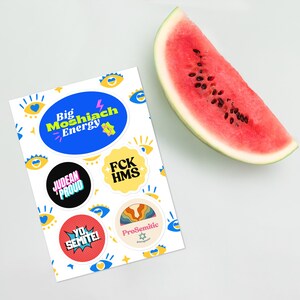 May include: A sheet of stickers with various designs and slogans, including "Big Moshiach Energy", "Judean Proud", "FCK HMS", "Yo Semite!" and "ProSemitic". The stickers are on a white background and there is a slice of watermelon in the background.