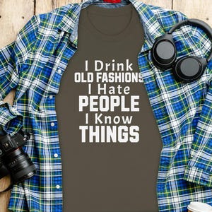 Könnte beinhalten: Ein dunkelolivgrünes T-Shirt mit dem weißen Text I DRINK OLD FASHIONS I HATE PEOPLE I KNOW THINGS, unter einem blau-grün-weiß karierten Hemd, ergänzt durch eine schwarze Kamera und einen schwarzen Over-Ear-Kopfhörer.