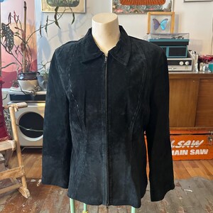 Peut inclure: Veste en daim noire avec un col et une fermeture éclair sur toute la longueur. La veste a des manches longues et une coupe ajustée, présentée sur un mannequin. La veste est de style classique, adaptée à diverses occasions.