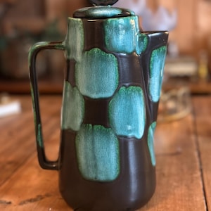 May include: A vintage ceramic coffee pot with a dark brown base and teal and green abstract pattern. The pot has a handle and a lid with a black accent. The coffee pot is sitting on a wooden surface.
