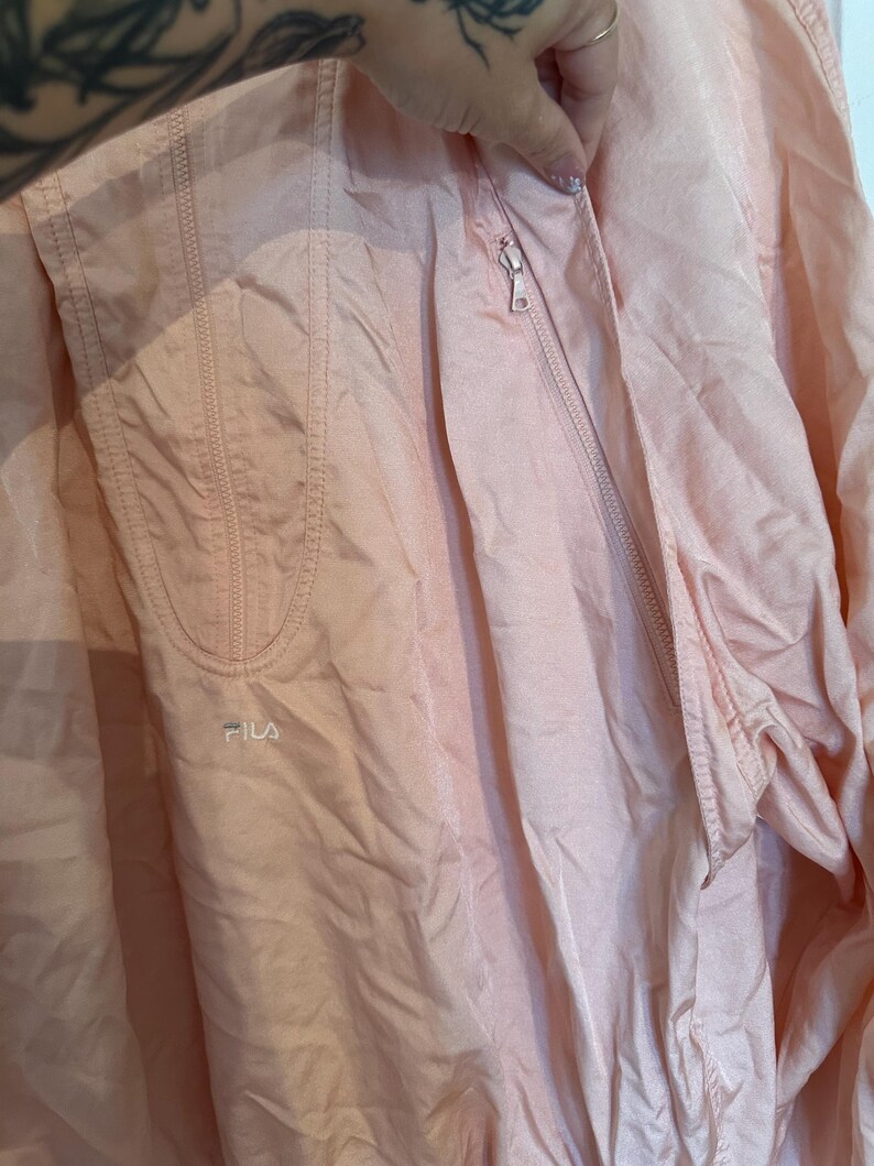 Puede incluir: Chaqueta cortavientos Fila rosa claro con cremallera. La chaqueta tiene un peque&ntilde;o logotipo de Fila en la parte delantera y un bolsillo en el lado izquierdo. La tela tiene un ligero brillo.