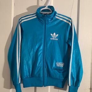May include: A blue Adidas track jacket with white stripes and the Adidas logo on the chest. The jacket has a zip fastening and a pocket on the left side. The text "adidas CHILE 62" is printed on the pocket.