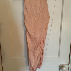 Puede incluir: Pantalones color melocot&oacute;n colgados de una percha de madera. Los pantalones tienen un tobillo fruncido y un corte holgado. La tela parece ligera y ligeramente arrugada. Los pantalones cuelgan de una puerta blanca.