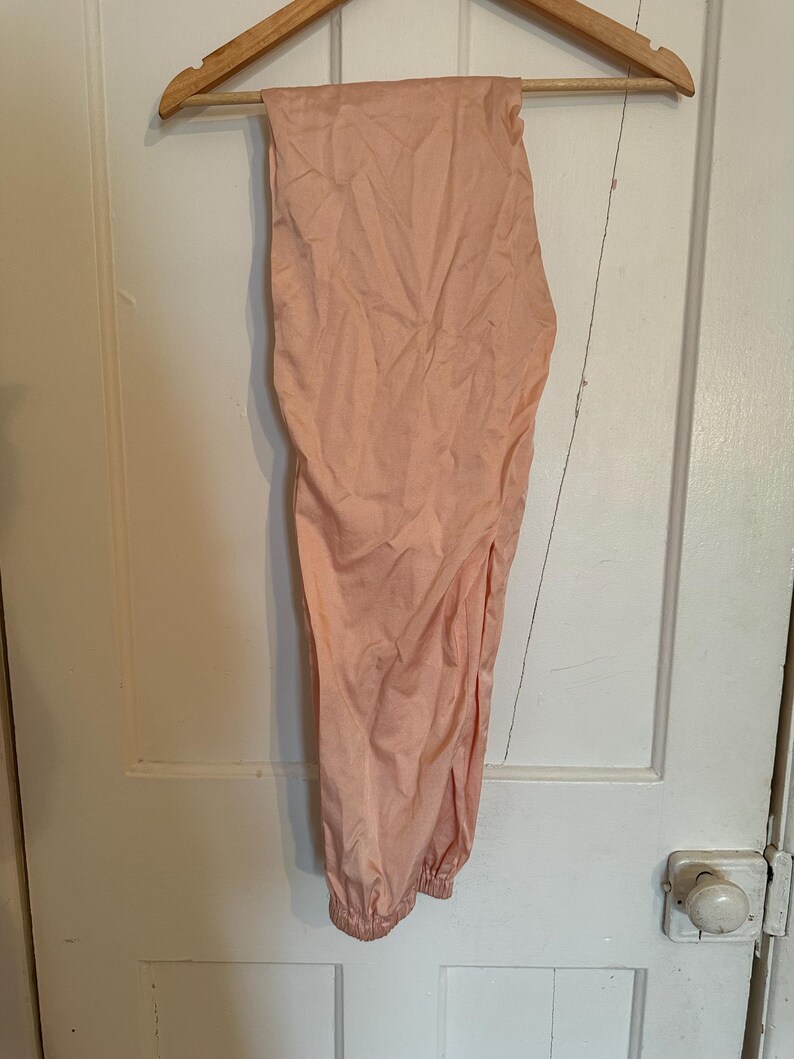 Puede incluir: Pantalones color melocot&oacute;n colgados de una percha de madera. Los pantalones tienen un tobillo fruncido y un corte holgado. La tela parece ligera y ligeramente arrugada. Los pantalones cuelgan de una puerta blanca.