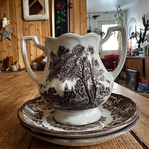 Peut inclure: Un service à crème et soucoupes en céramique blanche et marron. La crème est ornée d'un motif détaillé d'arbres et de maisons. L'ensemble est posé sur une table en bois. La crème a deux anses et un bord festonné.