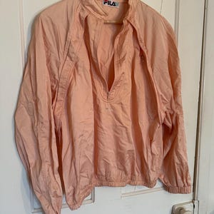 Puede incluir: Chaqueta cortavientos FILA de color melocot&oacute;n claro con cierre de media cremallera. La chaqueta tiene cuello alzado y pu&ntilde;os el&aacute;sticos. El logotipo de FILA es visible en el cuello y el pecho. La chaqueta est&aacute; colgada en una percha de madera.