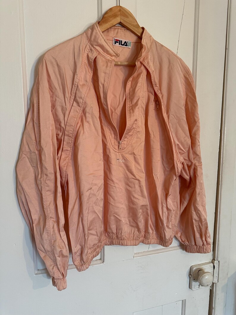 Puede incluir: Chaqueta cortavientos FILA de color melocot&oacute;n claro con cierre de media cremallera. La chaqueta tiene cuello alzado y pu&ntilde;os el&aacute;sticos. El logotipo de FILA es visible en el cuello y el pecho. La chaqueta est&aacute; colgada en una percha de madera.