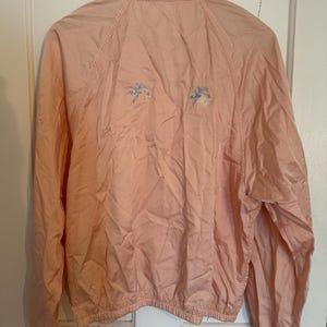 Puede incluir: Chaqueta rosa claro con cuello y mangas largas. La chaqueta tiene un dobladillo con cord&oacute;n y dos dise&ntilde;os florales bordados en la espalda. Cuelga de una percha de madera.