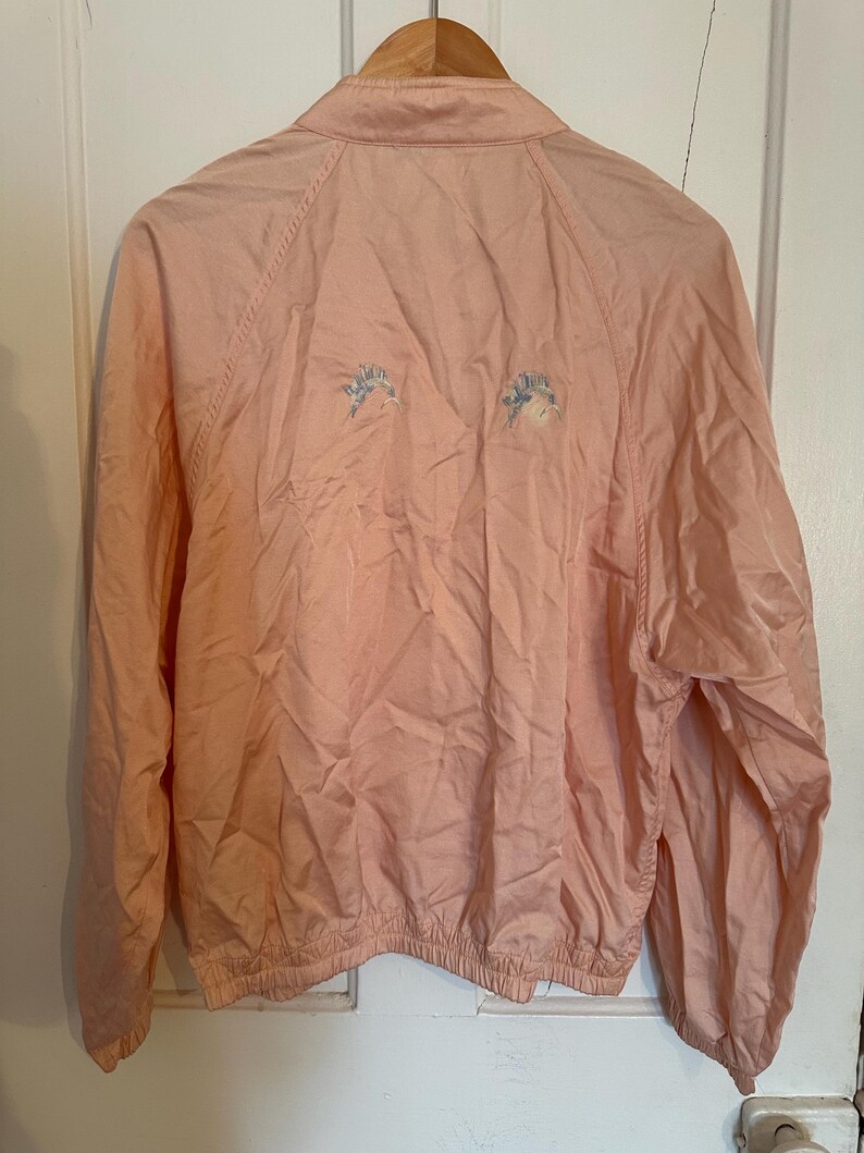 Puede incluir: Chaqueta rosa claro con cuello y mangas largas. La chaqueta tiene un dobladillo con cord&oacute;n y dos dise&ntilde;os florales bordados en la espalda. Cuelga de una percha de madera.