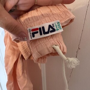 Puede incluir: Primer plano de una prenda de color melocot&oacute;n con un cord&oacute;n blanco. Se ve una etiqueta de Fila, con el logotipo de la marca en azul marino, rojo y blanco, con el texto "MADE IN ITALY". La etiqueta tambi&eacute;n incluye informaci&oacute;n sobre la talla.
