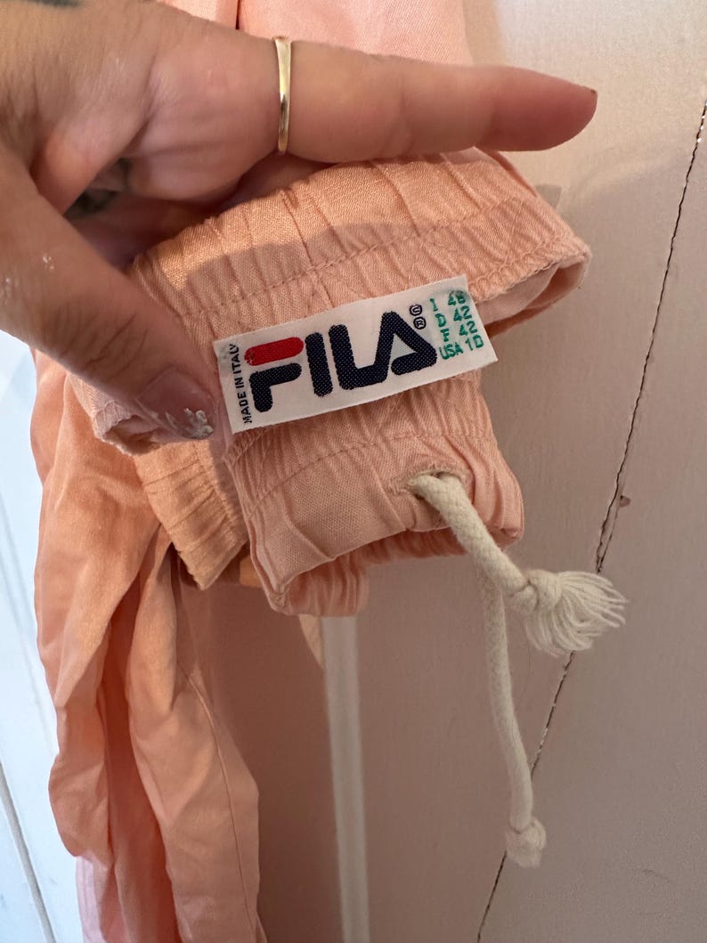 Puede incluir: Primer plano de una prenda de color melocot&oacute;n con un cord&oacute;n blanco. Se ve una etiqueta de Fila, con el logotipo de la marca en azul marino, rojo y blanco, con el texto "MADE IN ITALY". La etiqueta tambi&eacute;n incluye informaci&oacute;n sobre la talla.