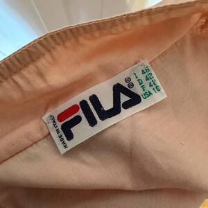 Puede incluir: Primer plano de una tela color melocot&oacute;n con una etiqueta Fila. La etiqueta es blanca con el logotipo de Fila en rojo y azul marino. La etiqueta tambi&eacute;n incluye informaci&oacute;n de tallas y "Made in Italy".