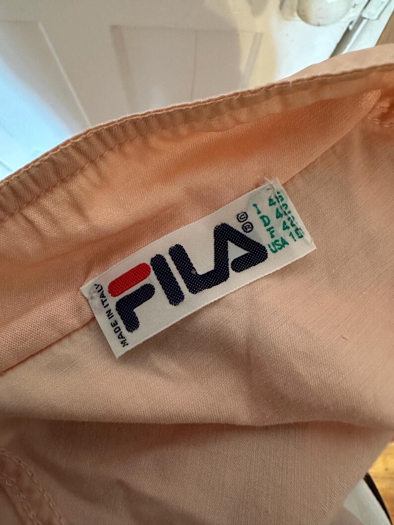 Puede incluir: Primer plano de una tela color melocot&oacute;n con una etiqueta Fila. La etiqueta es blanca con el logotipo de Fila en rojo y azul marino. La etiqueta tambi&eacute;n incluye informaci&oacute;n de tallas y "Made in Italy".