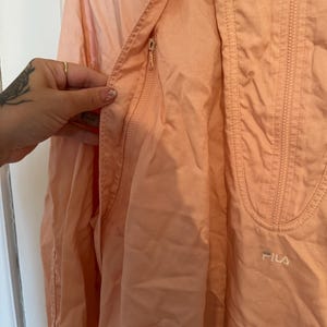Puede incluir: Chaqueta Fila de color melocot&oacute;n claro con cierre de cremallera y un peque&ntilde;o bolsillo. La chaqueta tiene una capucha con cord&oacute;n y el logotipo de Fila es visible en la parte inferior derecha. La chaqueta est&aacute; hecha de un material ligero.