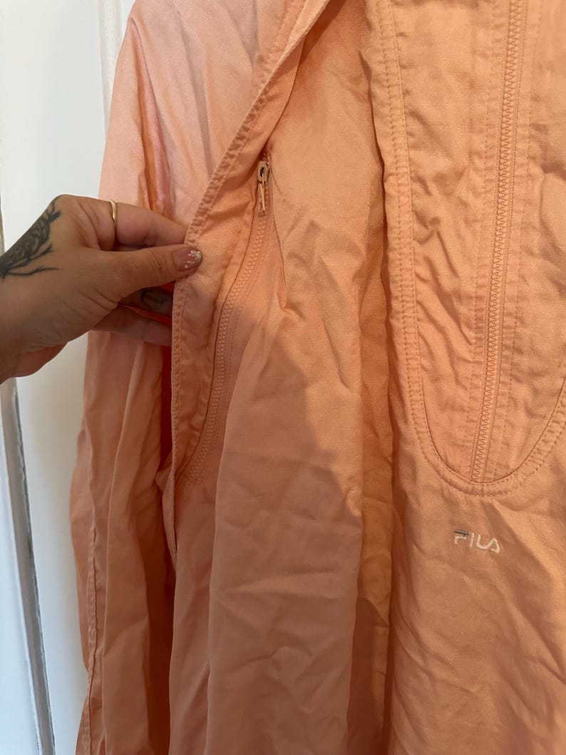 Puede incluir: Chaqueta Fila de color melocot&oacute;n claro con cierre de cremallera y un peque&ntilde;o bolsillo. La chaqueta tiene una capucha con cord&oacute;n y el logotipo de Fila es visible en la parte inferior derecha. La chaqueta est&aacute; hecha de un material ligero.