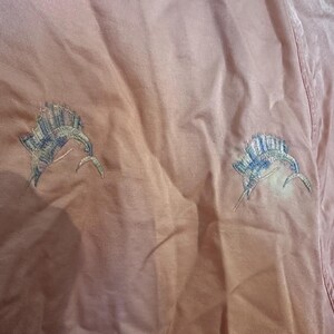 Puede incluir: Una camisa color melocot&oacute;n con dos dise&ntilde;os bordados de pez marl&iacute;n. Los peces est&aacute;n detallados con hilo azul, verde y blanco, y est&aacute;n colocados en la parte delantera de la camisa. La tela tiene un aspecto ligeramente texturizado.