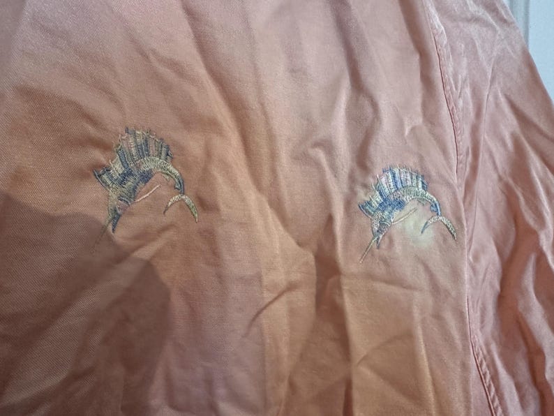 Puede incluir: Una camisa color melocot&oacute;n con dos dise&ntilde;os bordados de pez marl&iacute;n. Los peces est&aacute;n detallados con hilo azul, verde y blanco, y est&aacute;n colocados en la parte delantera de la camisa. La tela tiene un aspecto ligeramente texturizado.