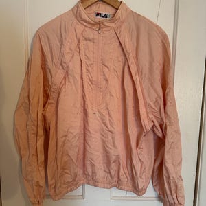 Puede incluir: Chaqueta FILA de color melocot&oacute;n claro con cuello alto y cierre de media cremallera. La chaqueta tiene mangas largas y cintura fruncida. El logotipo de FILA es visible en la cremallera y en el pecho. La chaqueta est&aacute; colgada en una percha de madera.