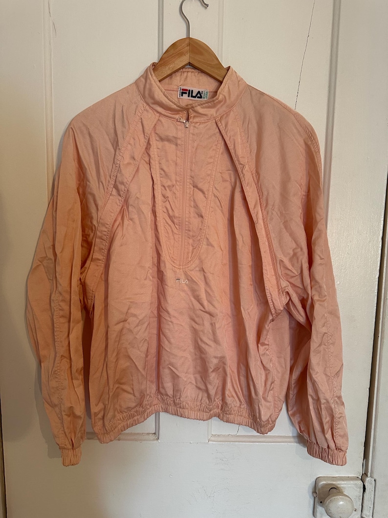 Puede incluir: Chaqueta FILA de color melocot&oacute;n claro con cuello alto y cierre de media cremallera. La chaqueta tiene mangas largas y cintura fruncida. El logotipo de FILA es visible en la cremallera y en el pecho. La chaqueta est&aacute; colgada en una percha de madera.
