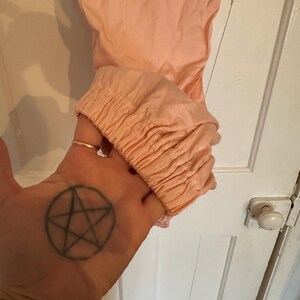Puede incluir: Una prenda de color melocot&oacute;n con un pu&ntilde;o fruncido. La tela parece ser un material ligero. La prenda est&aacute; sujeta por una mano con un tatuaje de pentagrama.