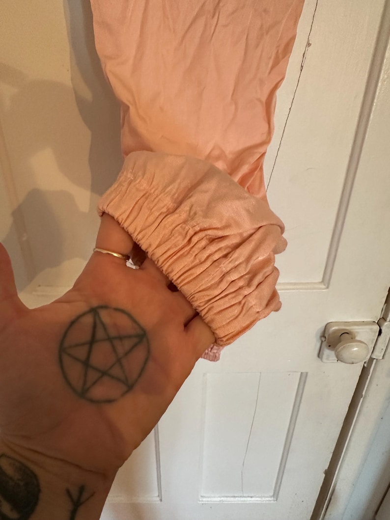 Puede incluir: Una prenda de color melocot&oacute;n con un pu&ntilde;o fruncido. La tela parece ser un material ligero. La prenda est&aacute; sujeta por una mano con un tatuaje de pentagrama.