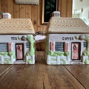 Peut inclure: Deux boîtes à thé et à café en céramique en forme de maison. Chacune a un toit beige, des murs blancs et une porte rose. Des vignes vertes et une clôture blanche décorent le devant. Les mots "TEA" et "COFFEE" sont écrits sur le devant.