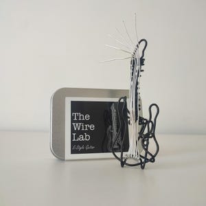 May include: A black and silver wire sculpture of an S-Style guitar. The sculpture is next to a metal tin with a black and white card that reads "The Wire Lab S-Style Guitar". The guitar sculpture has a unique, artistic design.