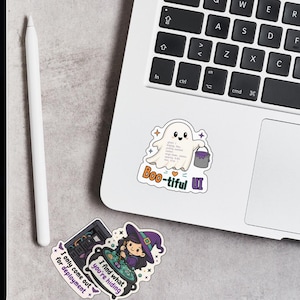May include: Two vinyl stickers on a laptop. One sticker features a white ghost holding a purple paint bucket with the text Boo-tiful UI. The second sticker shows a witch stirring a green cauldron with the text I find what you are hiding.