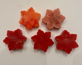 May include: Five snowflake-shaped wax melts in shades of red and orange. The melts are arranged in a row on a white surface.