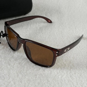 May include: Dark burgundy sunglasses with brown tinted lenses and a rectangular frame shape. The temples feature a silver-toned metallic logo emblem near the hinges, which are secured with two small silver rivets on each side of the front frame.