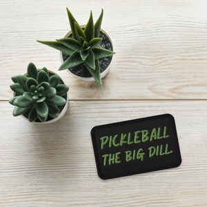 May include: Black patch with green text that reads "Pickleball The Big Dill" on a white wooden surface with two potted succulents.