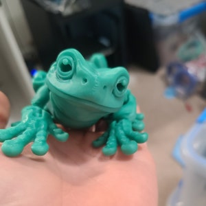 May include: A teal green frog figurine with large eyes and a wide smile. The frog is sitting on a hand.