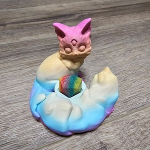 Puede incluir: Una figurita de gato de colores impresa en 3D con un dado de arcoíris en su boca. El gato está sentado sobre una base en forma de nube con un degradado de azul, blanco y rosa.