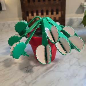 May include: A green and white plastic plant with a red pot. The plant has multiple leaves that are hinged and can be moved.