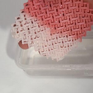 May include: A pink and white woven plastic mesh held above a clear plastic container. The mesh has a repeating pattern of interlocking shapes, with a gradient from pink to white. The container is partially filled with a clear liquid.