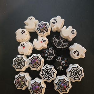 Könnte beinhalten: Eine Sammlung von Halloween-Anhängern mit Geistern, Fledermäusen und Spinnennetzen. Die Anhänger sind aus weißem und schwarzem Kunststoff und haben verschiedene Designs.