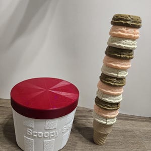 Könnte beinhalten: Ein weißer Behälter mit rotem Deckel und dem Text "Scoopy's" in einer strukturierten Schriftart. Daneben ein hoher Eisbecher mit mehreren Kugeln in verschiedenen Farben, darunter Braun, Rosa und Weiß, die übereinander gestapelt sind.