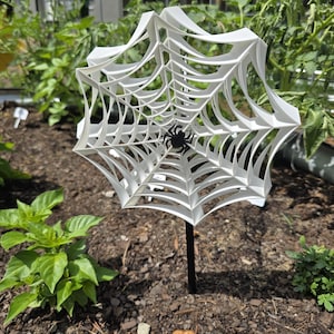 May include: A white, 3D-printed spider web garden stake with a black spider in the center. The web is mounted on a black stake, placed in a garden bed with green plants and dark soil. A decorative Halloween or spooky season garden accent.