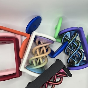May include: A collection of colorful, 3D-printed objects with a unique, geometric design. The objects feature a square frame with a central, intertwined spiral element. Colors include pink, orange, blue, green, purple, and black.