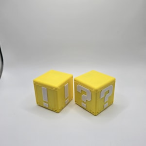 May include: Two yellow plastic blocks with white question marks and exclamation points on them. The blocks are from a popular video game.