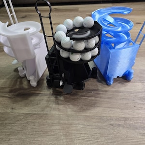 May include: Three golf ball storage and dispensing units in white, black, and blue. The black unit holds multiple golf balls on tiered shelves. The white and blue units have a spiral track design. All units have wheels and handles.