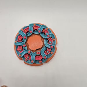 May include: A colorful, circular 3D-printed object with a central orange disc. The outer ring features a teal, wave-like design with small, red spheres. The object is likely a decorative piece or a unique toy.