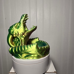 Könnte beinhalten: Eine grün-gelbe 3D-gedruckte Drachenskulptur mit offenem Maul, die auf einem weißen runden Sockel steht.