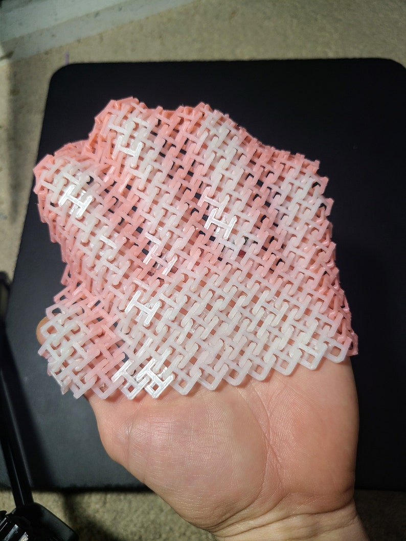 May include: A flexible, woven structure in shades of white and light pink, held in a hand. The pattern is a repeating series of interlocking shapes, creating a mesh-like appearance. The object appears to be 3D printed.