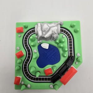 May include: A 3D printed model of a landscape featuring a green base with a black and white train track loop, a blue pond, a white mountain, several small green trees, and multiple buildings with red roofs and white walls arranged around the track.