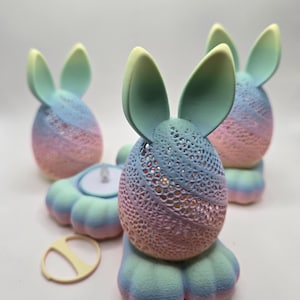May include: A collection of pastel-colored Easter bunny candle holders. Each bunny has a gradient of pink, blue, and yellow hues. The bunny's body is a perforated egg shape, and the ears are upright. The candle holders are on a white surface.