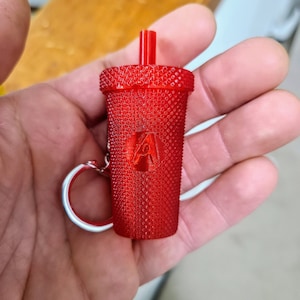 May include: A red plastic keychain shaped like a cup with a straw. The cup has a textured surface and a small silver ring attached to it.